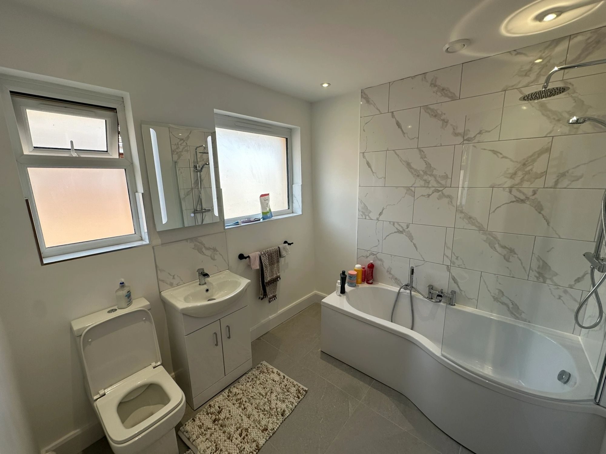 A finished bathroom with large marble-effect tiles, a shower over a curved bath, and a wall-hung basin unit.