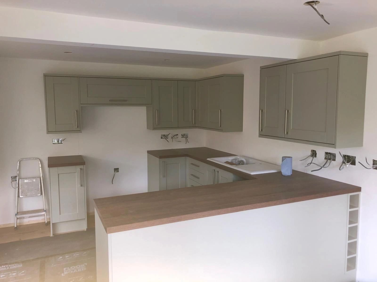 A kitchen part way through being fitted: sage green shaker units and worktops in place, sockets not yet dressed.